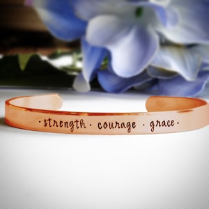 May include: A copper bracelet with the words "strength, courage, grace" engraved on it.