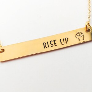 May include: Gold bar necklace with the words "Rise Up" and a raised fist symbol.