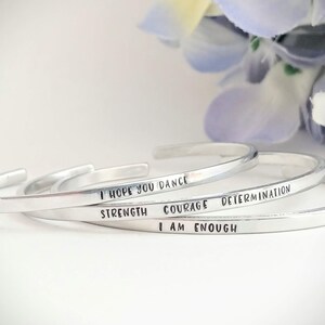 May include: Four silver bracelets stacked on top of each other. The bracelets are engraved with the words "I hope you dance", "strength", "courage", "determination", and "I am enough".