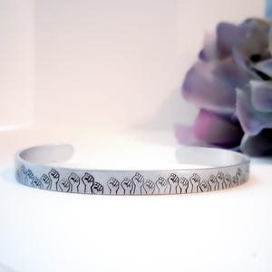 May include: Silver-toned cuff bracelet featuring a repeating pattern of raised fists. The open-ended bracelet is displayed on a white surface, with a blurred purple flower in the background. The design is a symbol of solidarity and empowerment.