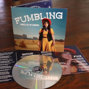 May include: A CD case with a colorful design featuring a woman wearing a yellow dress and a cowboy hat. The title of the album is "Fumbling" by Danielle Ate The Sandwich. The CD case is open and the CD is visible.