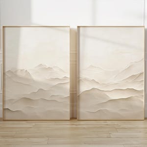 May include: Two framed landscape art prints. Each print features a serene mountain range in shades of beige and cream. The artwork is displayed in a room with light-colored walls and a wooden floor, creating a calming atmosphere.