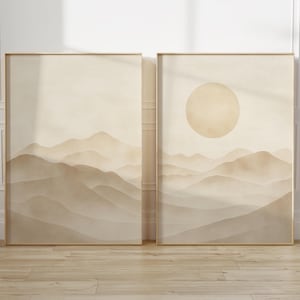 May include: Two framed landscape art prints. One features a mountain range, the other a sun over a mountain range. The artwork is in shades of beige and brown, with a minimalist aesthetic. The frames are gold.