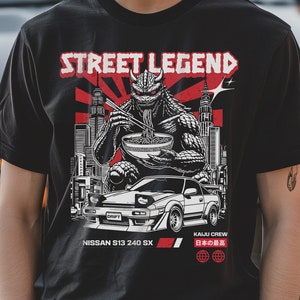 May include: Black t-shirt with a graphic featuring a monster eating noodles, a car, and the text "STREET LEGEND." The shirt also includes the text "NISSAN S13 240 SX" and "KAIJU CREW." The shirt is worn by a person.