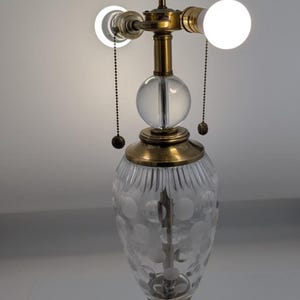 May include: A table lamp featuring a brass base and a clear glass body with etched polka dot patterns. The lamp has a central clear glass sphere, brass hardware, two light bulbs, and two pull chains for operation, standing on a square brass pedestal.