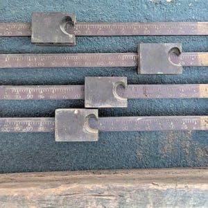 May include: An antique metal balance scale with a weathered, rustic appearance. The scale features a series of horizontal bars with measurement markings and sliding weights. The scale is designed to measure weight and has a vintage aesthetic.