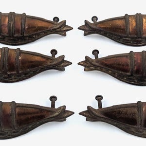 May include: A set of six metal drawer pulls featuring a curved, boat-like shape with a dark, weathered bronze finish. Each handle has a central raised section, pointed ends, and two attached mounting screws extending from the back of the hardware.