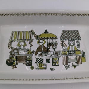 May include: Rectangular ceramic serving tray with a decorative border and handles. The tray features a whimsical illustration of three market stalls with people, food, and awnings in shades of green, brown, and white.
