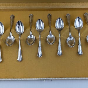 May include: A set of silver spoons arranged in a rectangular box with a yellow interior. The spoons have a reflective surface and ornate handles. The box has a cream-colored border.