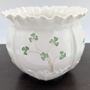May include: A white ceramic planter with a ruffled rim and a rounded body. The planter is decorated with green shamrock designs. The planter is suitable for indoor or outdoor use.