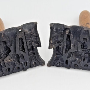 May include: Two antique black cast iron molds with wooden handles. The molds feature a detailed relief of figures on horseback and on foot, possibly depicting a historical scene. The handles are a light brown color.
