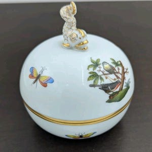 May include: A white, spherical trinket box with a gold rim and a dolphin-shaped lid handle. The box is decorated with colorful butterflies and birds perched on branches. The lid handle and rim have gold accents.