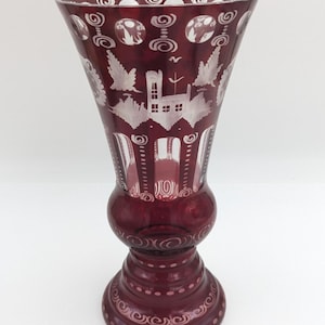 May include: A tall, ruby-red glass vase with intricate white etched designs. The vase features a flared top, a bulbous midsection, and a detailed base. The etched patterns include stylized birds, buildings, and swirling motifs, creating a decorative piece.