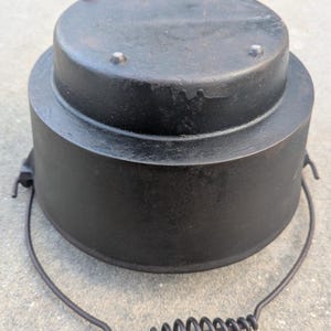 May include: A black cast iron Dutch oven with a lid and a wire handle. The lid has three small, raised bumps. The handle has a coiled spring grip. This cooking pot is designed for outdoor cooking.