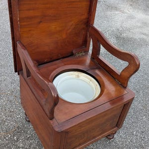 May include: Antique wooden commode chair with a hinged lid and armrests. The chair is made of dark wood and features a white, round basin. The chair has four small, rounded feet and a rectangular base.