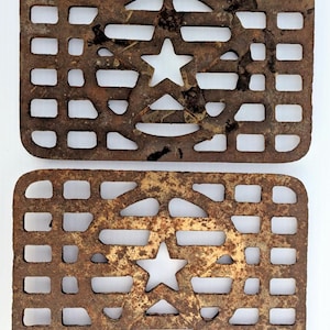 May include: Two rectangular, rusty metal grates. Each grate has a star-shaped cutout in the center and a grid of rectangular and curved openings. The metal has a weathered, aged appearance.