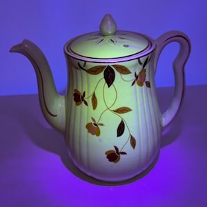 May include: A vintage cream-colored teapot with a brown and gold floral design. The teapot has a lid with a small hole in the top and a handle on the side.