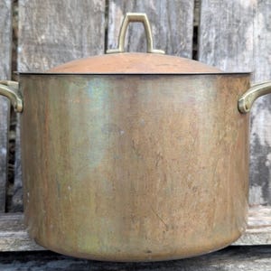 May include: A large, vintage copper pot with a lid and brass handles. The pot has a rounded body and a tarnished, aged appearance. The lid has a brass handle. The pot is designed for cooking.
