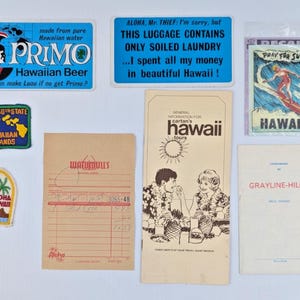 May include: A collection of vintage Hawaiian travel ephemera, including stickers, patches, and brochures. The items feature text such as "Primo Hawaiian Beer," "Aloha," and "Pray for Surf." The color palette includes blue, green, and tan.