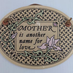 May include: Oval ceramic plaque with the text "MOTHER is another name for love." The plaque has a woven border design with green and pink floral accents. It includes a leather cord for hanging.