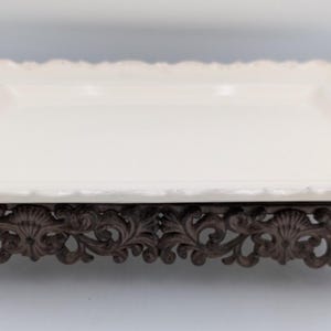 May include: A rectangular off-white ceramic serving tray with a scalloped edge, featuring two dark brown metal handles with ornate scrollwork. The tray is elevated by a matching dark brown metal base with intricate floral and shell detailing.