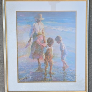May include: A framed painting depicts a serene beach scene. A person in a hat and dress stands with two children in the shallow water. The artwork features soft pastel colors, with a silver and gold frame.
