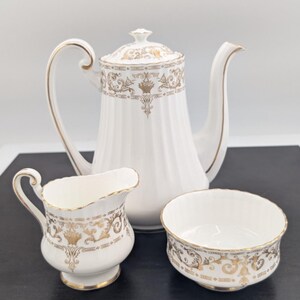 May include: A white porcelain tea set with gold accents. The set includes a tall coffee pot, a creamer, and a sugar bowl. The pieces are decorated with ornate gold patterns and have a classic, elegant design.