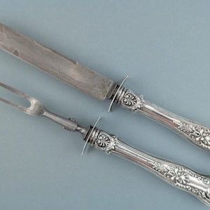 May include: Silver carving set with ornate handles. The set includes a knife and a two-tined fork. The handles feature a floral design. The knife blade is long and pointed. The fork has a curved shape.
