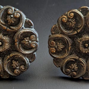 May include: Two antique bronze-colored cabinet knobs. Each knob features a central floral design surrounded by six ornate, swirling petal-like shapes. The knobs have a vintage, aged appearance, suggesting they are decorative hardware.