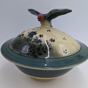 May include: Ceramic lidded bowl with a bird-shaped handle. The lid is decorated with blue and red flowers and green leaves. The bowl has a teal rim and base, and a cream-colored lid. The bird has a red head and dark wings.
