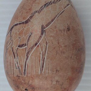 May include: A brown, egg-shaped stone carving featuring a giraffe. The giraffe's body is outlined in dark brown, with a white, geometric pattern. White lines depict the giraffe's legs and the grass it stands in.