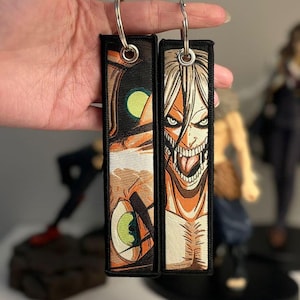 May include: Two rectangular keychains with anime-style illustrations. One shows a close-up of an eye with a green iris, the other a snarling face with exposed teeth. Both have black borders and silver key rings.