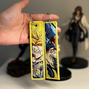 Puede incluir: Dos llaveros rectangulares amarillos con diseños bordados de personajes de anime. Un llavero presenta un personaje con cabello rubio puntiagudo, el otro tiene un personaje con armadura azul. Cada llavero tiene un anillo plateado.