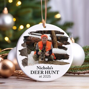 May include: A white, round Christmas ornament with a photo of a person in hunting gear holding a deer. The text "Nichola's DEER HUNT at 2025" is printed on the bottom. The ornament hangs from a gold string.