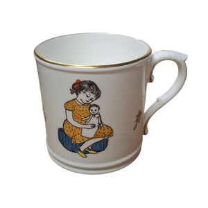 May include: White ceramic mug with a gold rim and a curved handle. The mug features a colorful illustration of a girl in a yellow dress holding a doll. The girl is sitting on a blue cushion. The mug is a standard size.