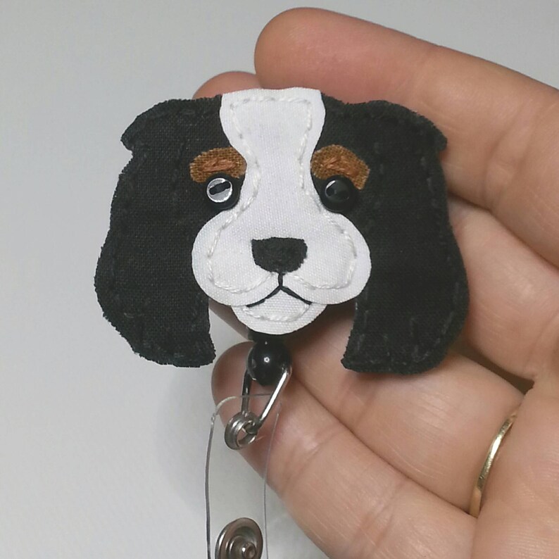 May include: A black and white felt Cavalier King Charles Spaniel dog badge reel with brown felt accents for the eyebrows. The dog has black eyes and a black nose. The badge reel has a black bead and a silver clip.