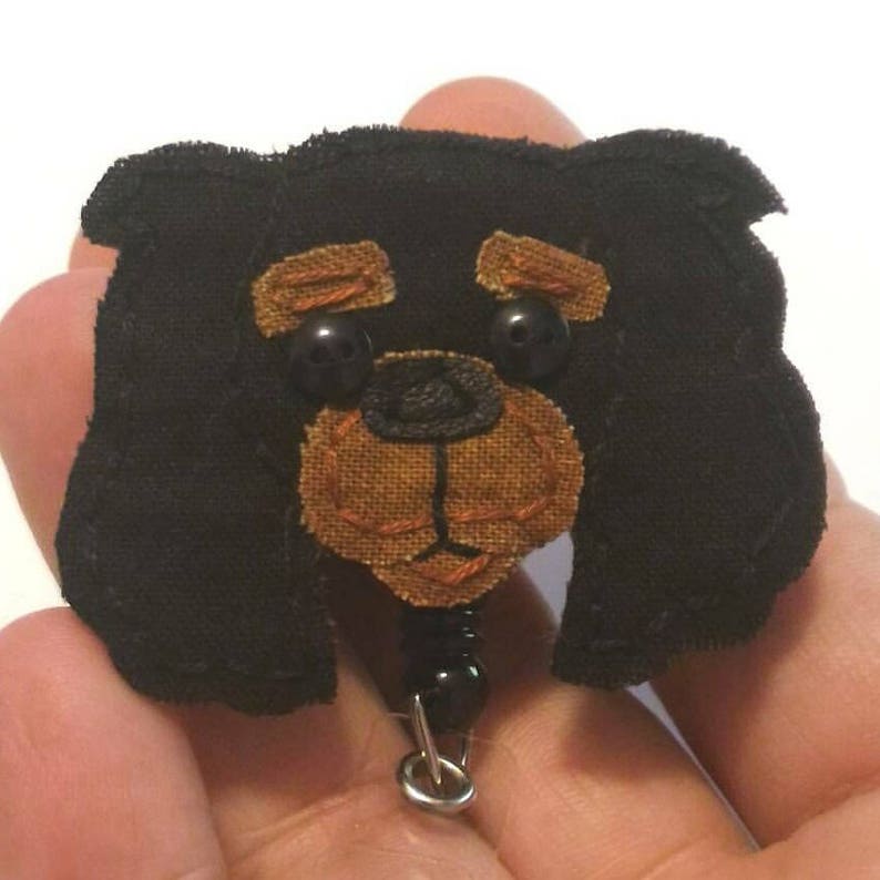 May include: Black fabric dog with brown felt face and black button eyes. The dog is attached to a black bead and a silver retractable badge reel.