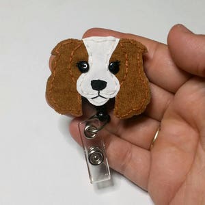 May include: A brown and white felt dog head with black eyes and a black nose, attached to a clear plastic retractable badge holder.