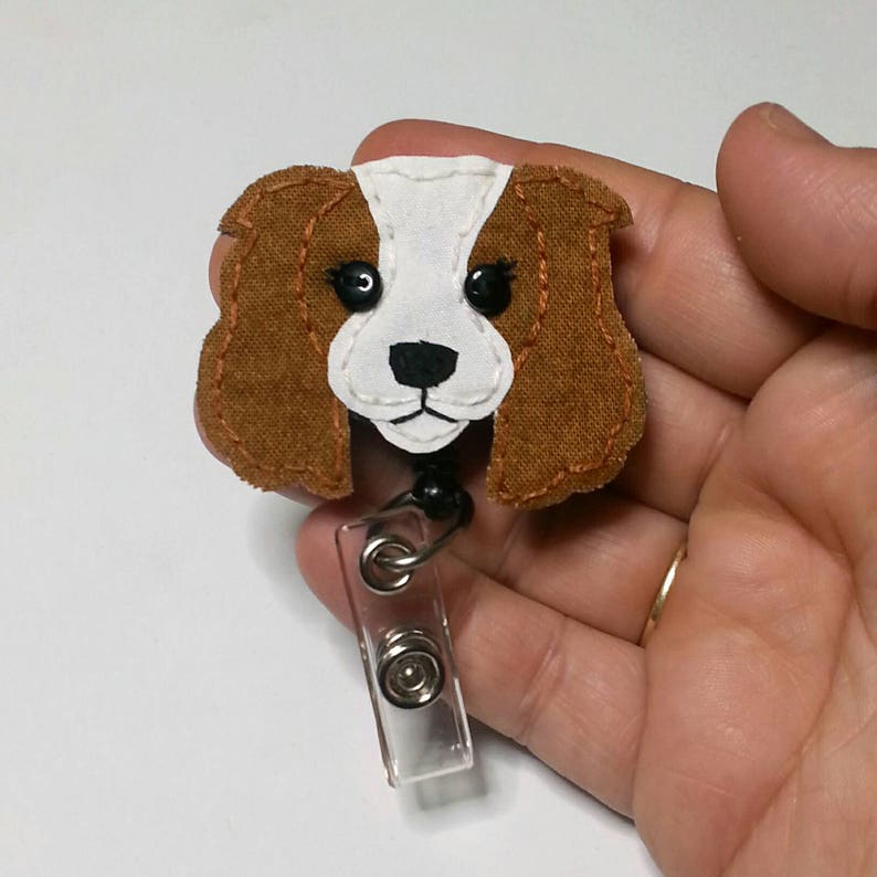 May include: A brown and white felt dog head with black eyes and a black nose, attached to a clear plastic retractable badge holder.
