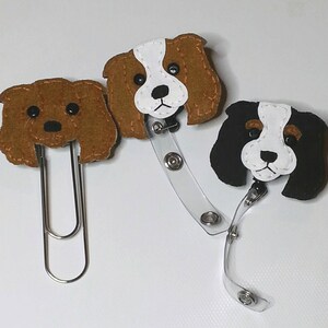 May include: Three felt dog badge holders. One brown dog with a clip, one brown and white dog with a retractable badge reel, and one black and white dog with a retractable badge reel.
