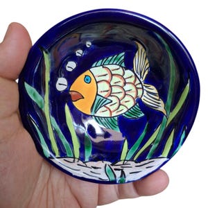 May include: A small blue ceramic dish with a hand-painted design of a yellow and green fish swimming in blue water with green seaweed and white bubbles.