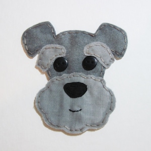 May include: A gray felt appliqué of a miniature schnauzer dog with black button eyes and a black felt nose. The dog has a stitched smile and is sewn with a white thread.