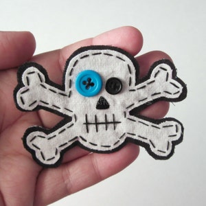 May include: A white fabric skull and crossbones with black stitching and blue and black button eyes.