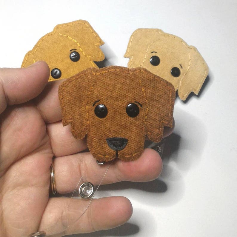 May include: Three felt dog badge holders with a retractable reel. The dogs are brown, tan, and light brown. The dogs have black eyes and black noses.