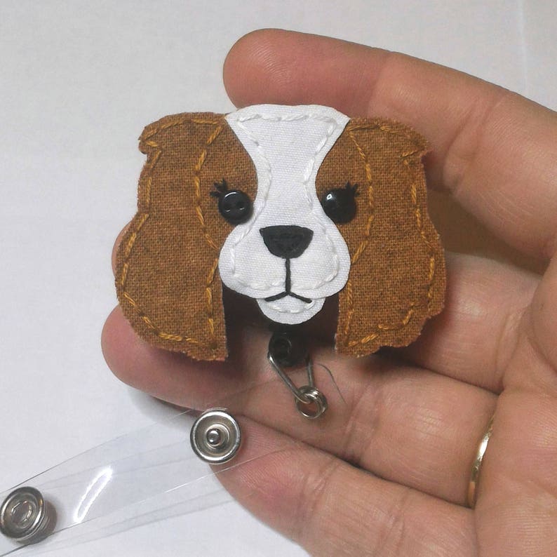 May include: A brown and white felt Cavalier King Charles Spaniel dog badge reel with black eyes and a white face. The reel is attached to a clear plastic badge holder.