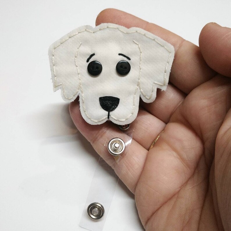May include: White fabric dog badge holder with black eyes and nose. The holder is attached to a clear retractable badge reel with silver metal clips.