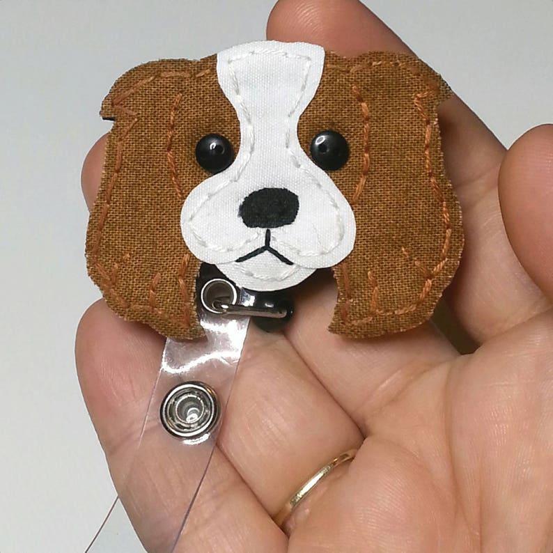 May include: A brown and white felt dog with black eyes and a black nose, attached to a clear retractable badge holder.