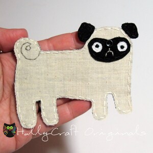 May include: A beige felt pug dog ornament with black felt ears and a black felt face. The pug has white felt eyes and a black felt nose.