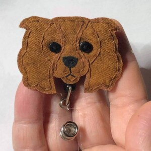 May include: A brown fabric Cavalier King Charles Spaniel dog head with black eyes and a black nose, attached to a clear plastic retractable badge holder with a silver clip.