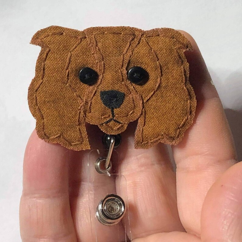 May include: A brown fabric Cavalier King Charles Spaniel dog head with black eyes and a black nose, attached to a clear plastic retractable badge holder with a silver clip.
